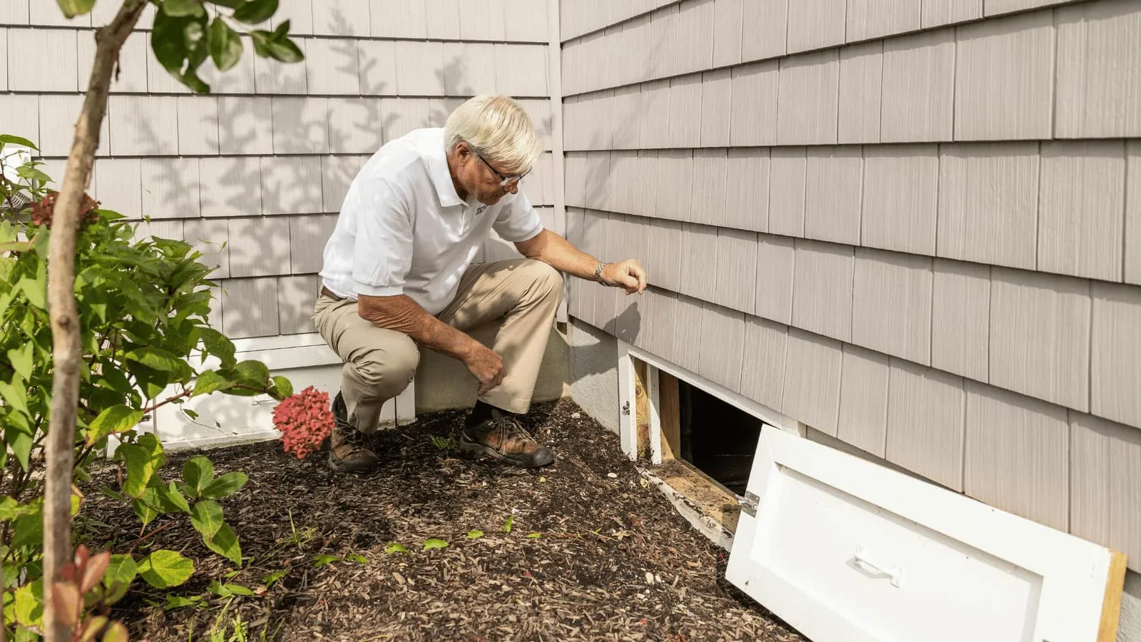 Inspector crouching to look at a foundation access panel beside the siding