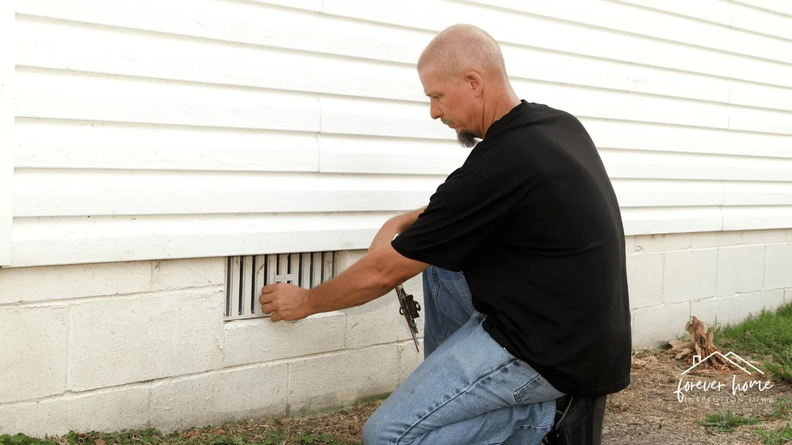 Inspector kneeling to check a foundation vent on a white-sided home