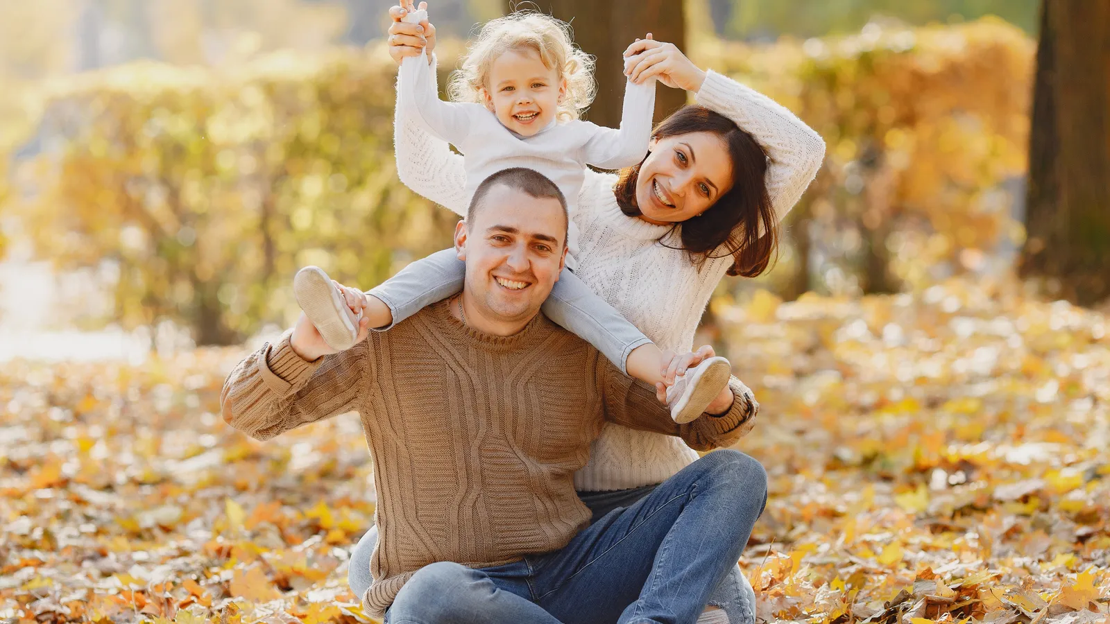 Father carrying a smiling child on his shoulders in fall leaves, with the mother beside them