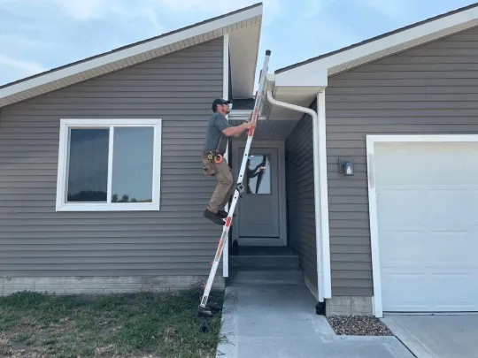Inspector on a ladder at the roofline of a grey ranch home